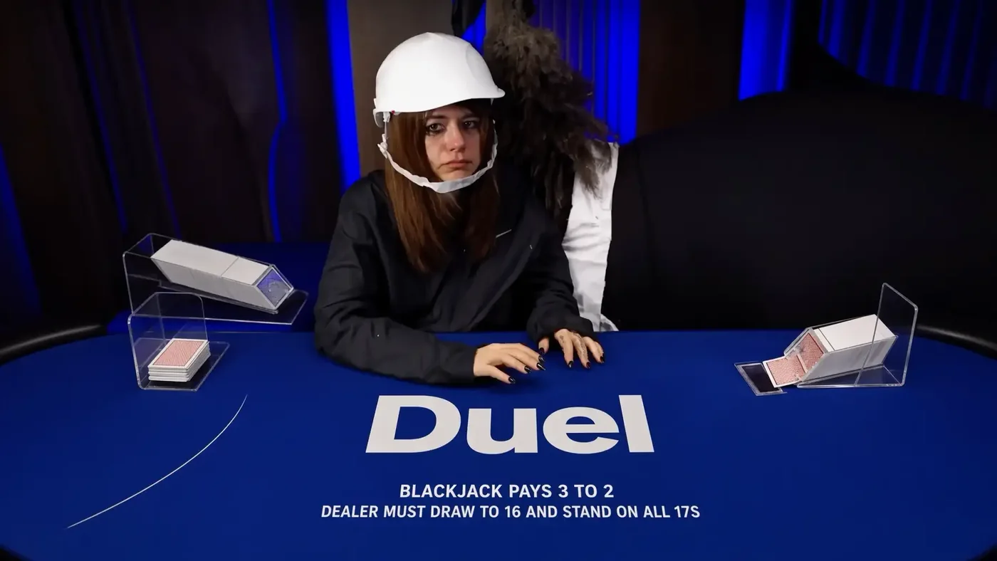 Annika dealing Duel Blackjack in a white hard hat and a dark zip jacket, hands on the blue felt that reads Duel, blackjack pays 3 to 2, dealer must draw to 16 and stand on all 17s