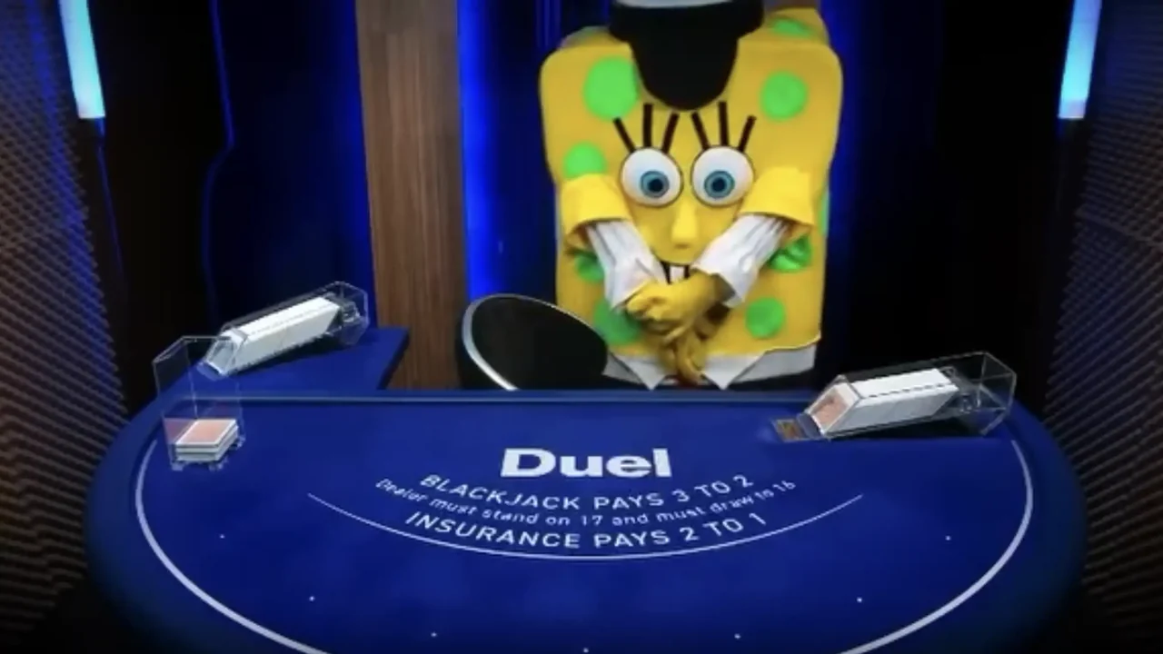 A yellow costume with green spots and oversized eyes standing at the dealer position of the Duel Blackjack table, hands folded, with the felt rules readable in front of it