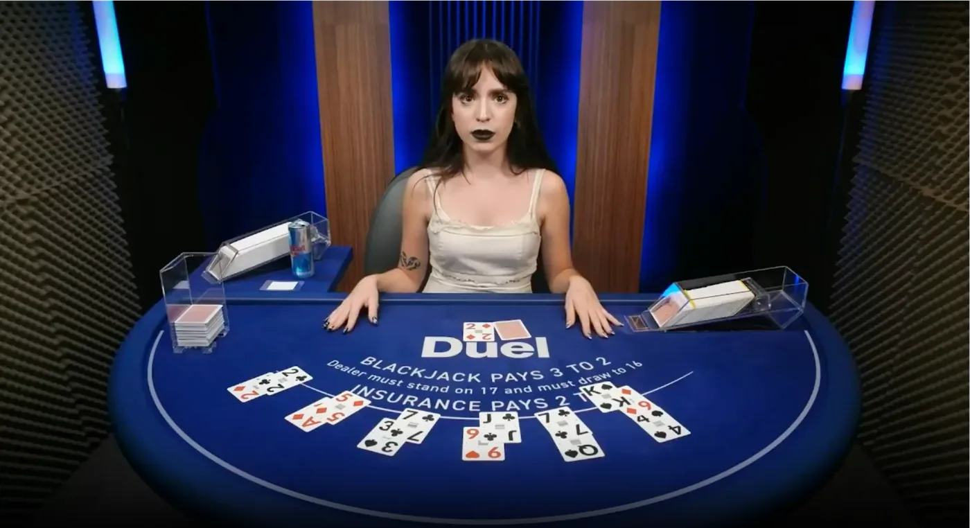 Nelli at the Duel Blackjack live table in a white top, both hands on the blue felt that reads Duel, blackjack pays 3 to 2, dealer must stand on 17 and must draw to 16